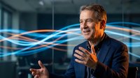 Middle-aged man with light skin tone and gray hair wearing dark blazer gesturing while speaking in modern office with blue digital wave graphics, representing business communication and technology innovation.