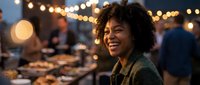 Young African American woman with curly hair smiling joyfully at outdoor evening market with warm bokeh lights and food vendors in background.