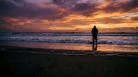 Silhouette of person standing on beach watching dramatic sunset over ocean waves with colorful sky reflecting on wet sand creating peaceful solitude moment.