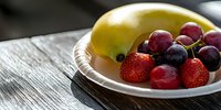 Fresh mixed fruits on white plate against rustic wooden background in natural lighting. Banana, strawberries and red grapes create healthy food composition.