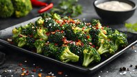 Fresh steamed broccoli florets garnished with red chili flakes and sesame seeds served on black plate against dark background, healthy vegetarian dish.