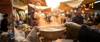 Person holding steaming bowl of soup at outdoor winter market with festive lights and crowd in background, holiday season celebration.