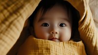 Asian baby wrapped in soft yellow muslin blanket looking at camera with bright curious eyes and gentle smile, warm golden tones create cozy atmosphere.