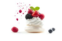 Fresh whipped cream meringue nest topped with ripe raspberries, blackberry and mint leaf on white background with berry juice splashes and blueberries.