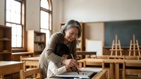Middle-aged Asian woman teacher preparing lesson materials at wooden desk in bright classroom with natural light from large windows and easels.