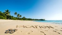 Tropical beach with word vacation written in sand, flip flops, palm trees, and clear blue sky on a sunny summer day.