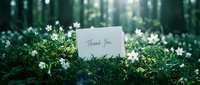 Thank you card resting on lush green moss surrounded by white wildflowers in a sunlit forest, expressing gratitude in nature.