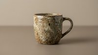 Rustic ceramic mug with speckled glaze finish and handle, isolated on neutral background. Suitable for tableware, kitchenware, and product design.