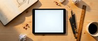Digital tablet with blank white screen on wooden desk surrounded by architectural tools, drafting supplies, coffee cup and crumpled papers for creative workspace mockup.