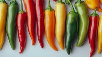Fresh colorful chili peppers in row including jalapeno, serrano, and habanero varieties on white background, showing different levels of heat and ripeness.