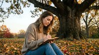 Young woman reading book under autumn tree during golden hour sunset in peaceful park setting with fallen leaves scattered around.
