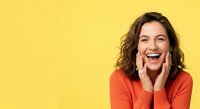 Young woman with wavy brunette hair and fair complexion wearing red shirt, smiling with hands on face against yellow background for advertising and marketing campaigns.
