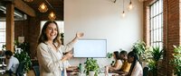 Professional businesswoman presenting to diverse team in modern office with exposed brick walls and plants for corporate training and workplace collaboration.