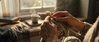 Elderly woman hands knitting with needles in cozy home interior with warm sunlight streaming through window creating peaceful domestic scene.