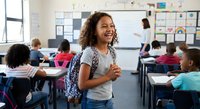 Happy mixed-race elementary school girl laughing in classroom with backpack, surrounded by diverse students at desks during school day.