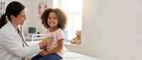 Female pediatrician examining happy African American girl child with stethoscope during medical checkup in bright clinic office for healthcare.