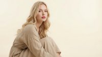 Young blonde Caucasian woman in beige oversized shirt sitting thoughtfully against neutral background for lifestyle and fashion content.