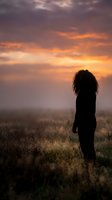 Silhouette of woman with curly hair standing in field during dramatic sunset with orange and purple sky clouds for contemplation and mindfulness concepts.