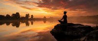 Person in silhouette meditating on rock by calm river at sunrise, reflecting on water with golden sky and trees. Wellness and nature concept.