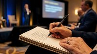 Professional businessman taking detailed notes during corporate presentation or conference with speaker and projection screen in background.