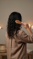 Woman with curly hair combing wet locks with wooden comb in cozy bathroom setting with candles for natural haircare routine and self-care wellness lifestyle.