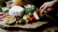 Mediterranean appetizer platter with fresh brie cheese, olives, cured meats, crusty bread, and colorful vegetables arranged on rustic wooden surface in moody lighting.