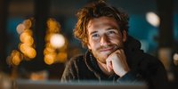 Young man with tousled hair smiling thoughtfully with hand on chin against blurred bokeh lights background, creating warm evening atmosphere.