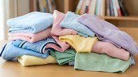 Pile of colorful folded baby clothes on wooden surface with bookshelf background. Soft pastel garments including onesies and shirts for newborn care.
