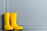 Bright yellow rubber rain boots against gray wall with classic molding, creating minimalist composition with strong visual contrast. For lifestyle and seasonal design.