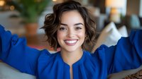 Young Hispanic woman with short wavy brown hair wearing bright blue blouse smiling warmly at camera with arms raised in joyful expression. Natural indoor lighting creates soft ambiance.