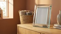 Empty glass photo frame on wooden dresser in warm cozy interior with wicker basket and vase, sunlight from window, mockup for design.