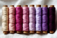 Colorful thread spools arranged in gradient from cream to deep purple, showing textile crafting supplies on white fabric background. Wooden spools display ombre color progression.