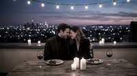 Romantic couple sharing an intimate dinner on a rooftop terrace with city lights, candles, and string lights glowing at dusk.