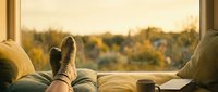 Cozy autumn afternoon at home, feet in warm wool socks resting on sofa by window with golden fall foliage view and coffee mug.