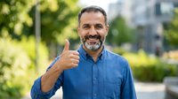 Middle-aged man with dark hair and beard, wearing blue denim shirt, giving thumbs up gesture outdoors with blurred green foliage and urban background. Suitable for business, success, approval, and lifestyle marketing.
