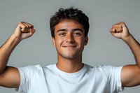 Young Hispanic man in white t-shirt expressing joy and triumph with raised fists and wide smile against gray background, showing positive emotions and success.