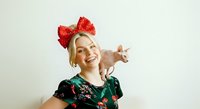 Young woman with blonde hair and fair complexion wearing red sequined bow headpiece and green floral top, holding small pig on shoulder against white background, studio portrait.