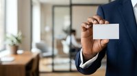 Professional man with dark skin tone wearing navy blazer and white shirt holds blank white business card in modern office environment with natural lighting and blurred background.