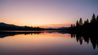 Serene lake at sunrise with evergreen forest silhouettes reflected in calm water, golden and purple sky, peaceful nature landscape for backgrounds and travel projects.