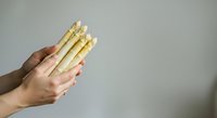 Cropped hand holding bundle of fresh white asparagus spears against neutral background, suitable for food styling, culinary content, and healthy eating promotions.