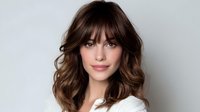 Young woman with wavy brown hair and bangs smiling at camera, wearing white top against light background, professional headshot style portrait.