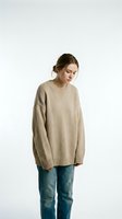 Young woman with brown hair in casual updo, fair complexion, wearing oversized beige knit sweater and blue denim jeans, standing against white background for fashion and lifestyle content.