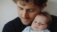 Tender moment between young Caucasian father in black shirt holding infant baby close, showing emotional connection and paternal love against light background.