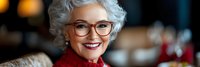 Senior Caucasian woman with curly gray hair and glasses smiling warmly in cozy indoor setting. Mature female portrait with joyful expression and red clothing.