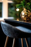 Modern black velvet bar stool with wooden legs against blurred Christmas tree background with golden ornament and bokeh lights creating festive atmosphere.