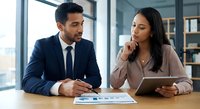 Professional business meeting with confident Hispanic man in navy suit and Asian woman in beige blazer discussing strategy with tablet and documents in modern office.