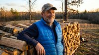 Smiling mature Caucasian lumberjack in blue vest and cap standing beside stacked logs at sunset in forest clearing during autumn logging operations.