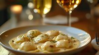 Homemade ravioli pasta with creamy sauce and herbs served on white plate in restaurant setting with wine glasses in background.
