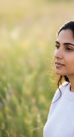 Young South Asian woman in white top gazing thoughtfully in sunlit green field, ideal for wellness, lifestyle, and portrait concepts.