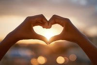 Hands forming heart shape silhouette against warm sunset sky with bokeh lights creating romantic atmosphere for love and relationship concepts.
